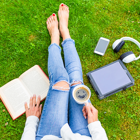 Top view of woman sitting on the green grass, relaxing with smartphone, headphones, tablet, book and coffee